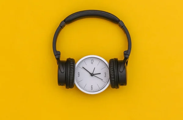 White clock with black stereo headphones on a yellow background. Top view