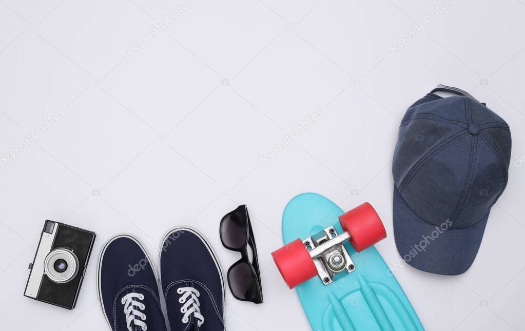 Flat lay composition of mini cruiser board, sneakers, cap, sunglasses and camera on white background. Youth, hipster outfit. Copy space. Top view