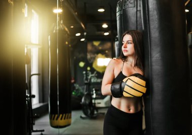 Boks salonundaki ağır çantanın yanında antrenman yaptıktan sonra yorgun ve zinde bir kadın dinleniyor. Kadın boksör idmandan sonra dinleniyor