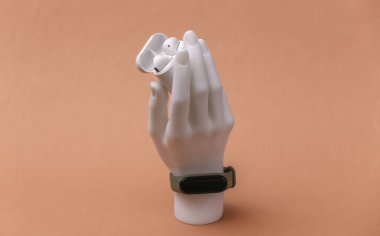 Beyaz el mankeni, kahverengi arka planda kablosuz kulaklık tutan akıllı bir bilezik ile ayakta duruyor. Modern aygıtlar