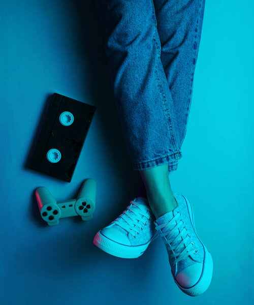 Retro wave still life. Women's legs in jeans sneakers, video tapes, gamepad. 