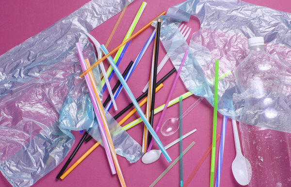 Концепция загрязнения окружающей среды. Целлофановый пакет, пластиковая бутылка, одноразовые вилки и ложки, коктейльные трубки на красном фоне. Вид сверху