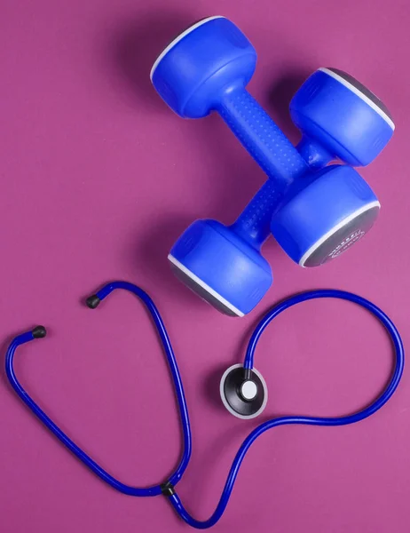 Healthy heart and strong muscles. Blue plastic dumbbells and stethoscope on purple background. Top view. Flat lay