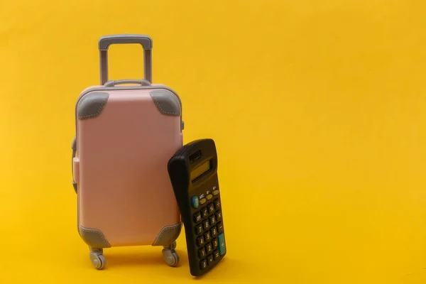 Travel expenses. Mini pink plastic travel suitcase with calculator on yellow background. Minimal style