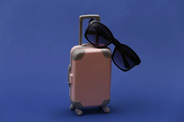 Travel or trip concept. Mini plastic travel suitcase with sunglasses on classic blue background. Color 2020