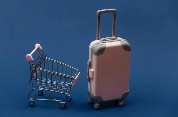 Travel and shopping concept. Mini plastic travel suitcase and supermarket trolley on classic blue background.