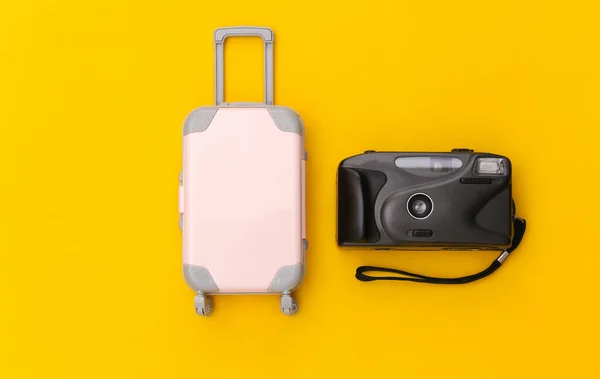 Travel flat lay. Mini plastic travel suitcase, camera on yellow background. Minimal style. Top view