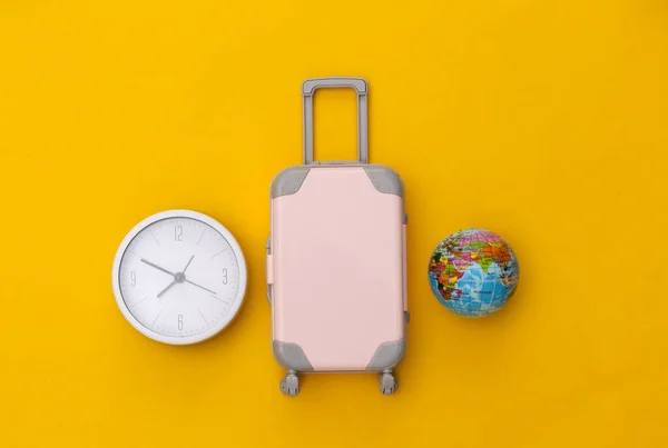 Time to travel. Mini plastic travel suitcase and clock, globe on yellow background. Top view. Flat lay