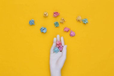 Beyaz manken el, sarı arka planda buruşmuş renkli kağıt topları tutuyor.