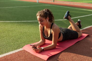 Neşeli sağlıklı kadın yoga minderinde dinleniyor ve stadyumda akıllı telefon kullanıyor.
