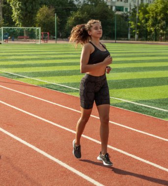 Spor giyimli zayıf bir kadın güneşli bir günde stadyumda koşuyor.