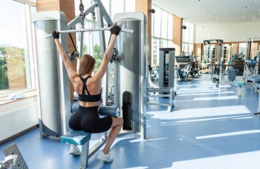 Güzel, genç, formda bir kadın spor salonundaki çekme makinesinde çalışıyor. Arka plan