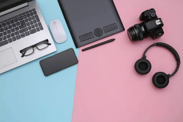 Workspace of photographer or retoucher. Laptop, graphic tablet, camera, smartphone and headphones on pink blue background. Top view.