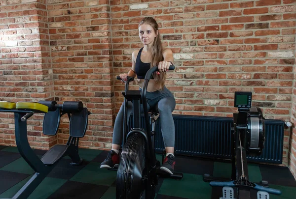 Attractive woman using exercise bike at gym. Fitness female using air bike for cardio workout at fitness gym