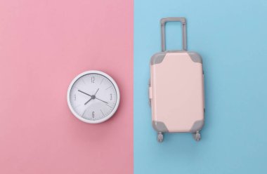 Seyahat zamanı. Mini plastik seyahat valizi ve pembe mavi pastel arka planda saat. Üst Manzara. Düz yatıyordu