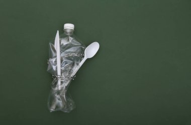 Buruşmuş plastik şişe, bıçak ve kaşık yeşil arka planda çelik bir zincire sarılı. Eko konsepti. Üst görünüm