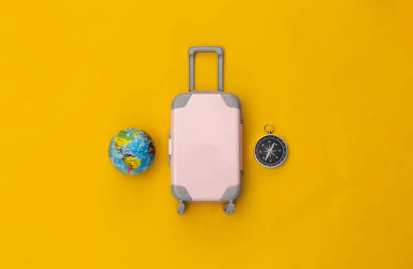 Travel flat lay. Mini plastic travel suitcase, globe and compass on yellow background. Minimal style. Top view