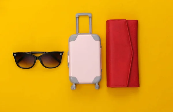 Travel flat lay. Mini plastic travel suitcase, red wallet and sunglasses on yellow background. Minimal style. Top view