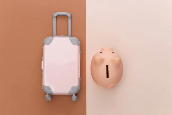 Travel minimalism. Mini plastic travel suitcase and piggy bank on beige brown background. Minimal style. Top view, flat lay