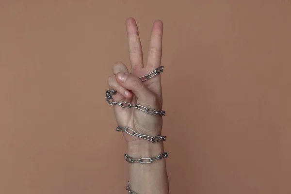 V fingers gesture, hand wrapped in a metal chain on a brown background. Suppression of freedom or imprisonment