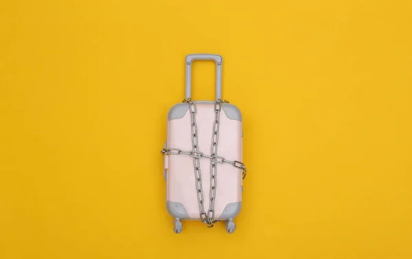 Mini travel baggage wrapped in steel chain on yellow background. Travel ban. Top view