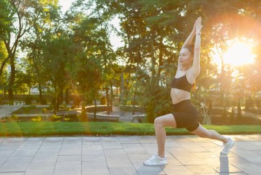 Savaşçı pozu. Genç, sıska kadın, gün doğumunda parkta yoga yapıyor.