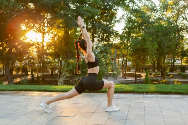 Savaşçı pozu. Genç, sıska kadın, gün doğumunda parkta yoga yapıyor.