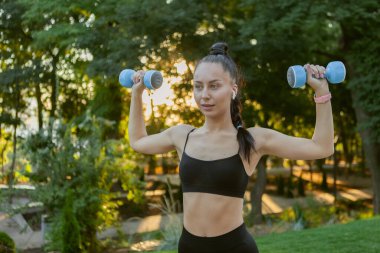 Çekici beyaz tenli fit esmer kadın güneş doğarken parkta elinde halterlerle omuz kaslarını çalıştırıyor.