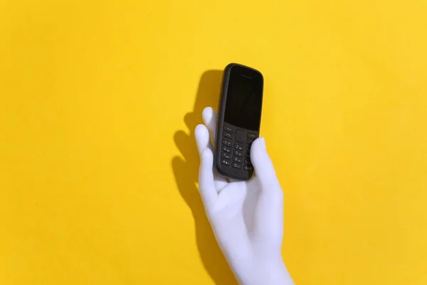 Mannequin\'s hand holds a push-button telephone on a yellow background. Top view