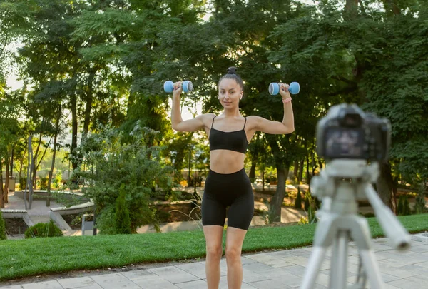 Online fitness trainer. Young attractive fit woman shows how to do exercise with dumbbells, filming blog on camera outdoors in the morning. Sports blog, fitness vlog