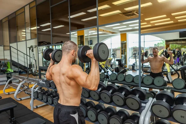 在现代体育馆里 赤身裸体的肌肉男一边做哑铃运动一边照着镜子 — 图库照片