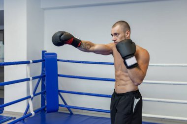 Çıplak gövdeli kaslı bir adam ringde boks eldivenleriyle el yumruğu atıyor.