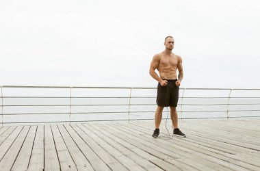 Kabartmalı kaslı adam sahilde ip atlıyor.