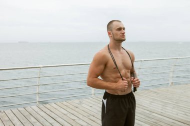 Kabartmalı, çıplak gövdeli, kumsalda boynunda ip olan kaslı bir adam.