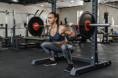 Spor salonunda omuzlarında ağır bir halterle çömelme egzersizi yapan genç bir kadın.