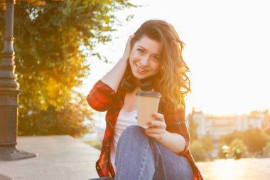 Çekici, gülümseyen öğrenci bir kadının portresi. Genç hippi kız gün doğumunda şehirde bir fincan kahve tutuyor.