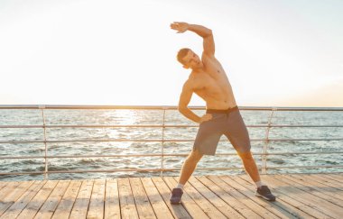 Çıplak vücutlu kaslı yakışıklı adam güneş doğmadan önce plajda egzersiz yapıyor.