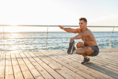 Çıplak gövdeli kaslı yakışıklı adam gün doğumunda sahilde tek ayak egzersizi yapıyor.