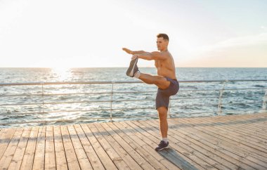 Çıplak vücutlu kaslı yakışıklı adam güneş doğmadan önce plajda egzersiz yapıyor.