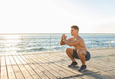 Çıplak vücutlu kaslı yakışıklı adam gün doğumunda sahilde çömelme egzersizi yapıyor.