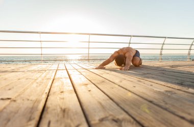 Asana yogası yapan kaslı bir adam kumsalda güneş doğarken kaslarını germek için poz verir.