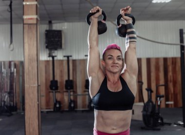 Kaslı, olağanüstü bayan sporcu, kısa pembe saçlı, kafasının üstünde ağır kettlebell 'ler var. Modern spor salonunda işlevsel, çapraz eğitim.