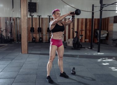 Kısa pembe saçlı, kaslı, olağanüstü bayan sporcu ağır kettlebell 'lerle sallanıyor. Modern spor salonunda işlevsel, çapraz eğitim.