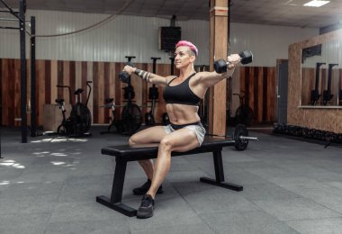 Genç, atletik, güçlü bir kadın spor salonundaki bir bankta otururken elinde dambıllarla omuz kaslarını çalıştırıyor. Vücut geliştirme ve Fitness