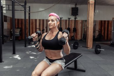 Spor salonundaki bir bankta otururken, kaslı atletik kadın elinde dambıllarla bisepsleri çalıştırıyor..