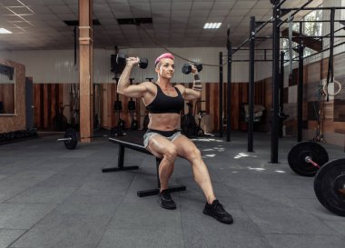 Atletik güçlü kadın, çapraz spor salonundaki bir bankta otururken elinde dambıllarla omuz kaslarını çalıştırıyor. Vücut geliştirme ve Fitness