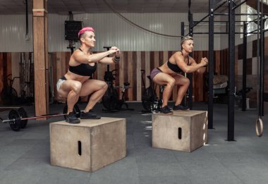 Spor kıyafetli atletik kadınlar spor salonunda tahta kutunun üzerinde dikilirken çömelip bacaklarını çalıştırıyorlar. Çapraz eğitim