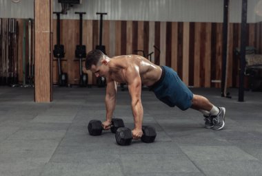 Spor salonunda dambıllarla şınav çeken kaslı güçlü bir adam.