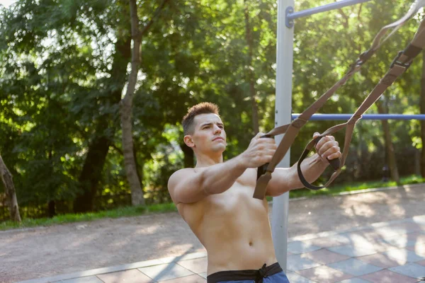 年轻英俊的男子赤身裸体的躯干在室外运动场上训练与桁架阻力系统 健康生活方式概念 — 图库照片