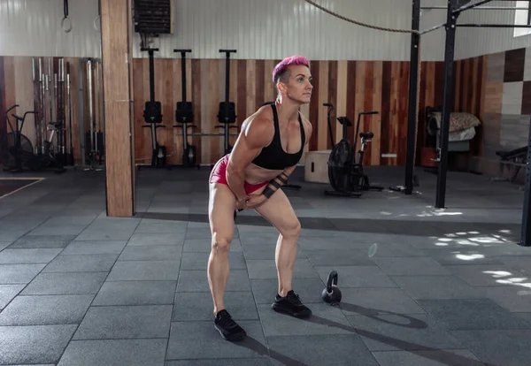 Kısa pembe saçlı, kaslı, olağanüstü bayan sporcu ağır kettlebell 'lerle sallanıyor. Modern spor salonunda işlevsel, çapraz eğitim.